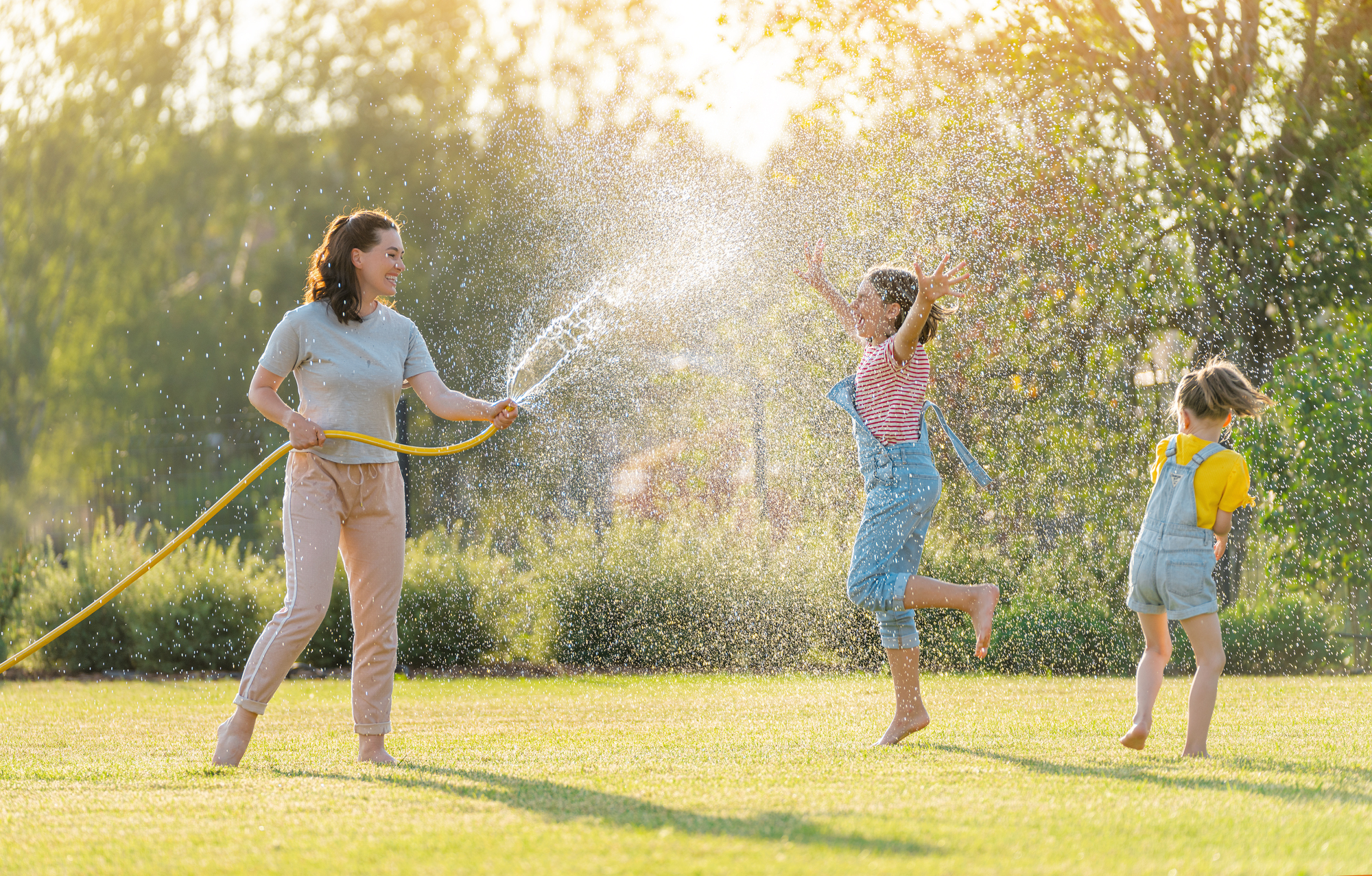 Happy family playing in backyard. Mother sprinkling her kids in hot summer day.